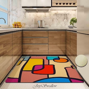 Può includere: Un tappeto geometrico astratto colorato con un contorno nero e una superficie testurizzata. Il tappeto è in una cucina con armadi in legno e un piano di lavoro bianco.