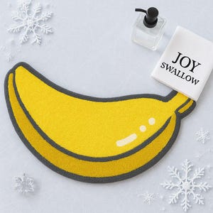 May include: A yellow banana-shaped bath mat with a grey border. A white towel with the words "JOY SWALLOW" is next to the mat. A clear soap dispenser and snowflake decorations are also in the image.