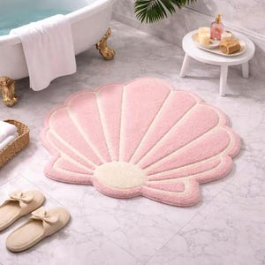 May include: A pink seashell-shaped bath mat with white accents. The mat is made of a soft, plush material and is placed on a marble tile floor. The mat is near a white bathtub and a pair of beige slippers.