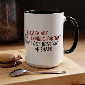 May include: White ceramic coffee mug with a black rim and handle. The mug has a red and black inscription that reads "Blessed are the flexible for they don't get bent out of shape."
