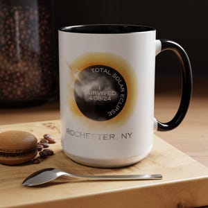 May include: White ceramic mug with a black rim and handle. The mug features a graphic of a total solar eclipse with the text "I survived Total Solar Eclipse 4/08/24 Rochester, NY".