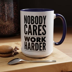 May include: White ceramic mug with a navy blue interior and handle. The mug features the text "NOBODY CARES WORK HARDER" in bold black letters. A spoon, coffee beans, and a macaron are on a wooden surface.