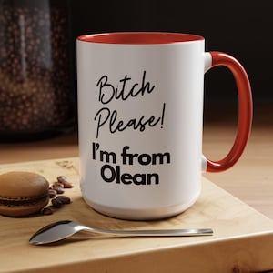 May include: White ceramic mug with a red interior and handle. The mug features the text "Bitch Please! I'm from Olean" in black script. A macaron, coffee beans, and a spoon are on a wooden surface.