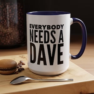 May include: White ceramic mug with a navy blue interior and handle. The mug features the text "EVERYBODY NEEDS A DAVE" in bold black letters. A spoon, a macaron, and coffee beans are on a wooden cutting board.