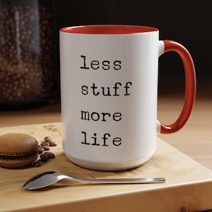 May include: White ceramic mug with a red rim and handle. The mug has the text "less stuff more life" printed in black.