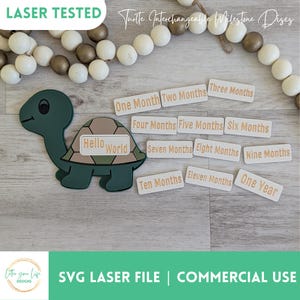 May include: A green turtle with a brown shell and the words "Hello World" on it. There are 12 white rectangular labels with gold text that say "One Month", "Two Months", "Three Months", "Four Months", "Five Months", "Six Months", "Seven Months", "Eight Months", "Nine Months", "Ten Months", "Eleven Months", and "One Year".
