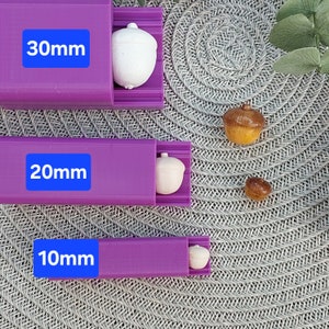 Könnte beinhalten: Drei violette Kunststoffformen in verschiedenen Größen: 30 mm, 20 mm und 10 mm. Jede Form hat eine kleine eichelförmige Vertiefung. Auf einer grauen, strukturierten Oberfläche befinden sich außerdem drei kleine eichelförmige Objekte, eines in jeder Größe.