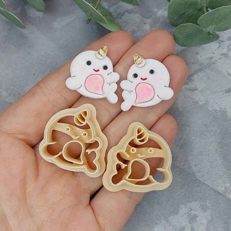 May include: Two white fondant narwhal cookie cutters and two plastic narwhal cookie cutters. The fondant cutters are decorated with gold glitter and pink hearts. The plastic cutters are a light brown color.
