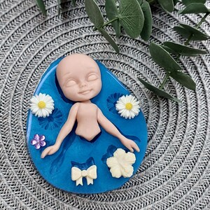 Pode incluir: Um molde de silicone azul com uma cavidade em forma de boneca, cercada por cavidades menores para flores e um la&ccedil;o. A boneca tem os olhos fechados e uma express&atilde;o doce.