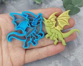 Könnte beinhalten: Ein blauer, drachenförmiger Ausstecher neben einer grünen Drachenfigur. Der Ausstecher ist eine detaillierte Umrisszeichnung eines Drachen mit Flügeln, während die Figur ein solider, dreidimensionaler Drache ist. Der Drache hat einen langen Schwanz und detaillierte Flügel.