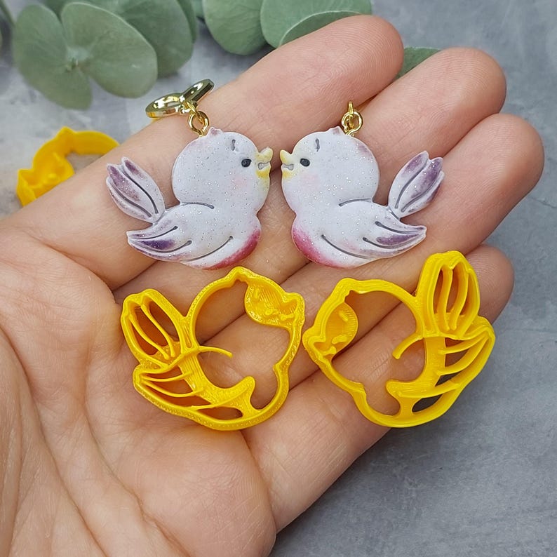 May include: A pair of white bird earrings with gold accents and a pair of yellow bird-shaped cookie cutters.