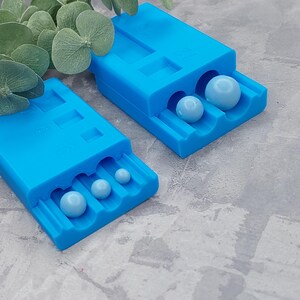 Peut inclure: Deux moules en plastique bleu pour la fabrication de perles rondes. Les moules ont des cavités de différentes tailles pour la fabrication de perles de différentes tailles. Les moules sont remplis de petites perles rondes bleu clair.