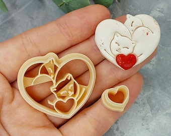 May include: Two cookie cutters in the shape of hearts. One cutter features two cats curled up inside the heart, the other cutter is a simple heart shape.