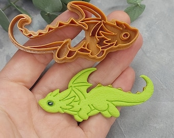 Könnte beinhalten: Ein grüner Drachenkeks mit detailliertem Design und einem passenden hellbraunen Ausstecher. Der Drache hat Flügel, einen Schwanz und einen freundlichen Ausdruck. Der Ausstecher ist aus Kunststoff.