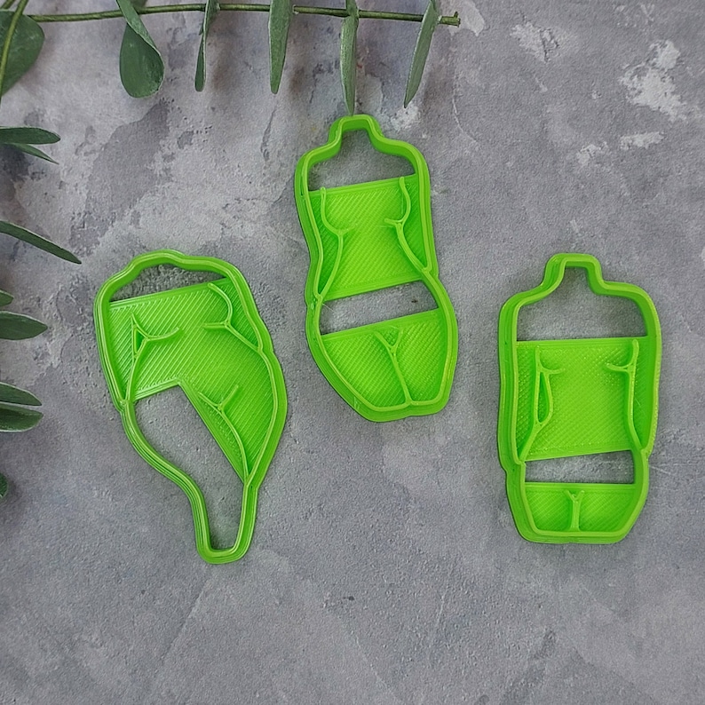 May include: Three bright green plastic cookie cutters in the shape of a human body. The cutters are arranged on a gray surface, with a sprig of greenery in the background. The cutters are designed to cut out the shape of a torso and legs.
