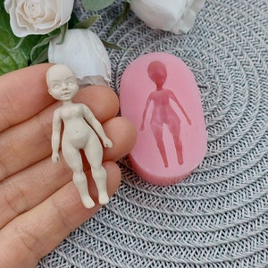 Op de afbeelding: Een roze siliconen mal voor het maken van een klein poppenfiguur. De mal is gevormd als een persoon met armen, benen en een hoofd. Naast de mal is een klein, wit poppenfiguur te zien.