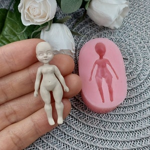 Op de afbeelding: Een siliconen mal voor het maken van een klein poppenfiguur. De mal is roze en heeft een gedetailleerde omtrek van een pop met armen, benen en een hoofd. Naast de mal is een klein, ongeverfd poppenfiguur te zien.