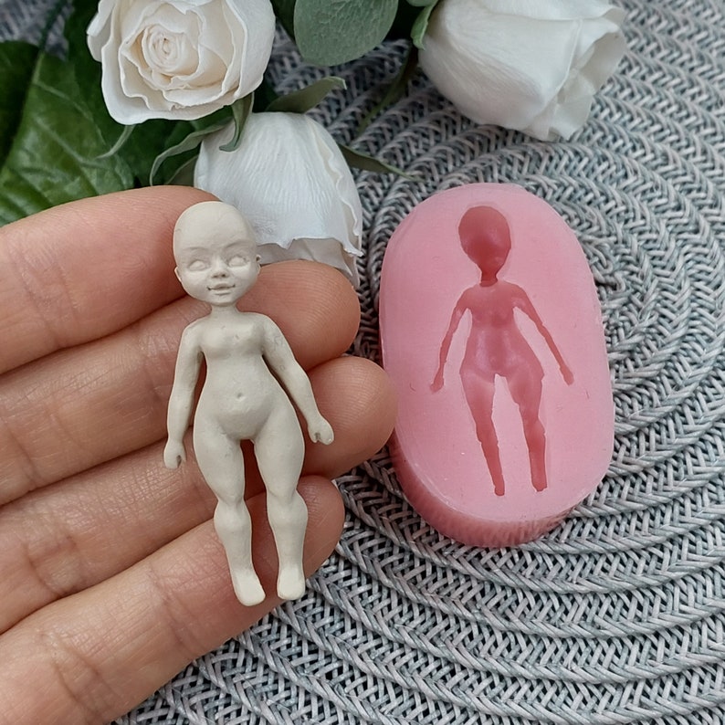 Op de afbeelding: Een siliconen mal voor het maken van een klein poppenfiguur. De mal is roze en heeft een gedetailleerde omtrek van een pop met armen, benen en een hoofd. Naast de mal is een klein, ongeverfd poppenfiguur te zien.