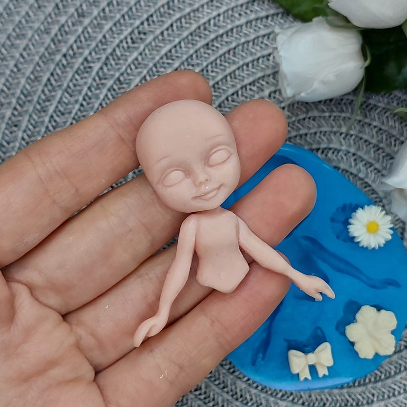 Pode incluir: Um molde de silicone para fazer uma pequena figura de boneca. O molde &eacute; azul e tem um desenho de uma boneca com os bra&ccedil;os estendidos, um la&ccedil;o, uma flor e uma pequena flor.