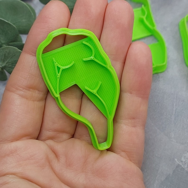 May include: A bright green leaf-shaped cookie cutter. The cutter has a detailed vein pattern and is held in a hand. Other cutters are visible in the background. The cutter is made of plastic.
