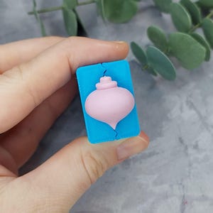 Könnte beinhalten: Eine blaue Silikonform zum Herstellen eines rosafarbenen Weihnachtsbaumanhängers. Die Form ist wie ein traditioneller Weihnachtsbaumanhänger mit einer Spitze geformt.