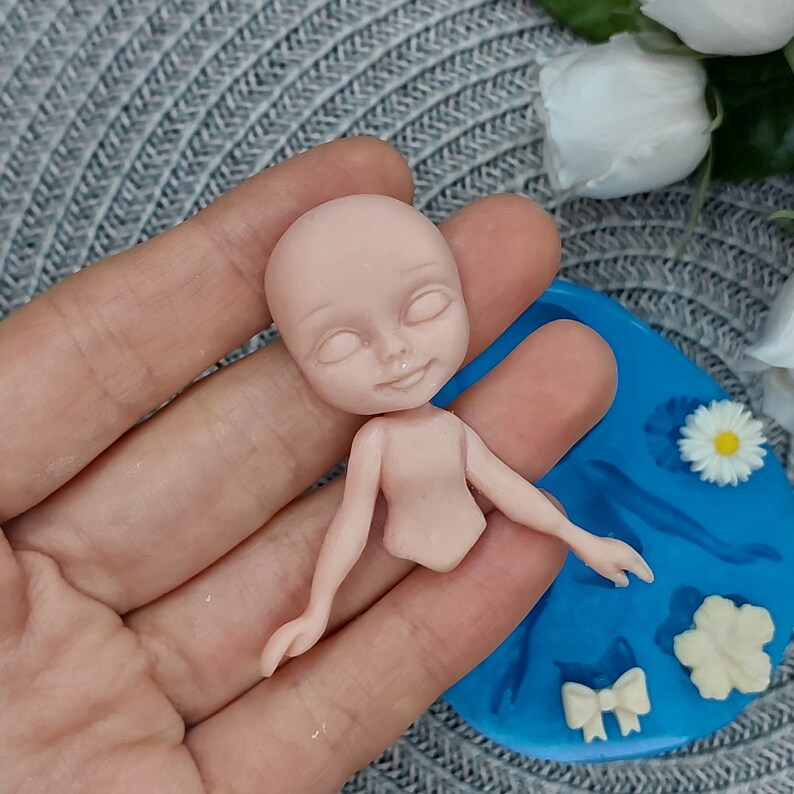 Pode incluir: Um molde de silicone para fazer uma pequena figura de boneca. O molde &eacute; azul e apresenta um corpo de boneca, um la&ccedil;o, uma flor e uma pequena flor. Uma figura de boneca parcialmente terminada &eacute; mostrada na m&atilde;o de uma pessoa.