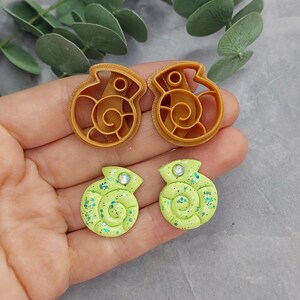 May include: Two tan seashell-shaped cookie cutters and two green seashell-shaped earrings with glitter and a small gem. The earrings are a light green colour with a spiral design. The cookie cutters are made of plastic.