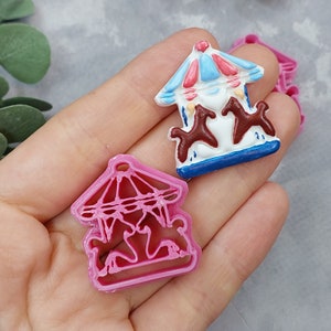Puede incluir: Dos cortadores de galletas de plástico rosa en forma de carrusel. Un cortador es un contorno detallado de un carrusel con caballos. El otro cortador es un carrusel sólido con caballos y un techo azul y blanco.