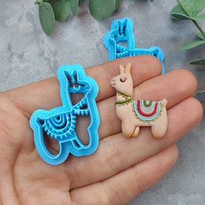 May include: Two llama cookie cutters, one blue and one beige. The blue cutter is detailed with a patterned design. The beige cutter is decorated with colorful stripes and gold accents.