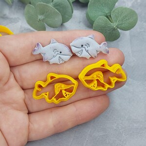 Op de afbeelding: Twee witte keramische haaienkoekjesvormen en twee gele plastic haaienkoekjesvormen. De koekjesvormen liggen op een hand.