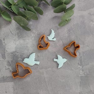 May include: A collection of bird-shaped cookie cutters and cut-out biscuits. The cutters are orange, and the biscuits are light blue. Eucalyptus leaves are in the background. Suitable for baking and crafts.