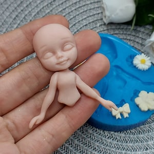Pode incluir: Uma pequena boneca de porcelana n&atilde;o pintada com os olhos fechados e uma boca sorridente. A boneca est&aacute; sendo segurada em uma m&atilde;o, e um molde de silicone azul com v&aacute;rias formas &eacute; vis&iacute;vel no fundo.