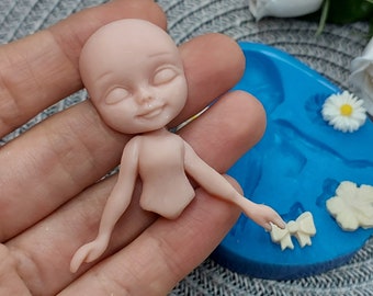 Puede incluir: Una pequeña muñeca de porcelana sin pintar con los ojos cerrados y una boca sonriente. La muñeca está siendo sostenida en una mano, y un molde de silicona azul con varias formas es visible en el fondo.