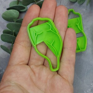 May include: Two bright green leaf-shaped cookie cutters. One is held in a hand, the other is in the background. The cutters have detailed vein patterns and are made of plastic. The background includes eucalyptus leaves.