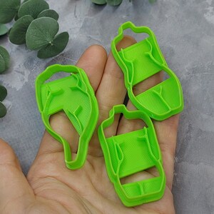 May include: Three bright green plastic cookie cutters in the shape of a bottle. The cutters are held in a hand, with a gray background and green leaves in the background. The cutters have a handle and a rectangular body.
