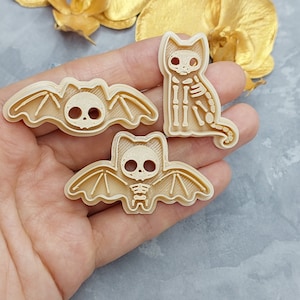 Puede incluir: Tres cortadores de galletas, un gato, un murciélago y un murciélago, todos con diseños de esqueleto. Los cortadores están hechos de plástico beige.