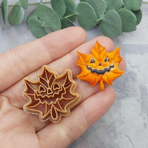 May include: A pumpkin-orange maple leaf-shaped Halloween decoration with a carved jack-o'-lantern face. Next to it is a brown plastic cookie cutter in the same shape. The leaf has a stem and is held in a hand.