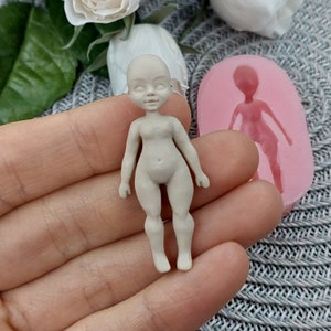 Op de afbeelding: Een kleine, ongeverfde, witte porseleinen pop met een gedetailleerd gezicht en lichaam. De pop wordt in een hand gehouden, en een roze siliconen mal van de pop is zichtbaar op de achtergrond.