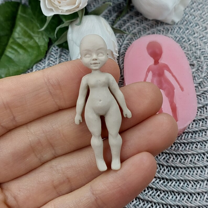 Op de afbeelding: Een kleine, ongeverfde, witte porseleinen pop met een gedetailleerd gezicht en lichaam. De pop wordt in een hand gehouden, en een roze siliconen mal van de pop is zichtbaar op de achtergrond.