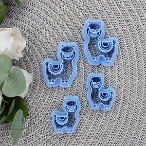 May include: Four blue plastic llama cookie cutters on a gray textured surface. The cookie cutters are different sizes and have a simple design.