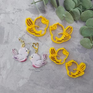May include: A set of four yellow plastic bird-shaped cookie cutters and two pairs of earrings featuring a pink bird design. The earrings have a gold heart charm.