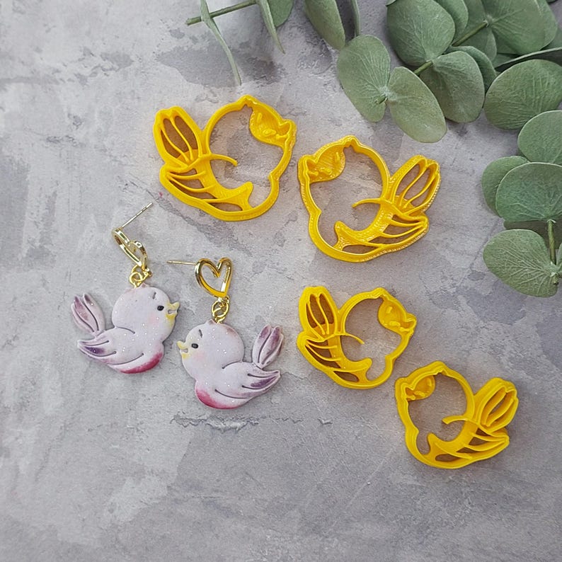 May include: A set of four yellow plastic bird-shaped cookie cutters and two pairs of earrings featuring a pink bird design. The earrings have a gold heart charm.