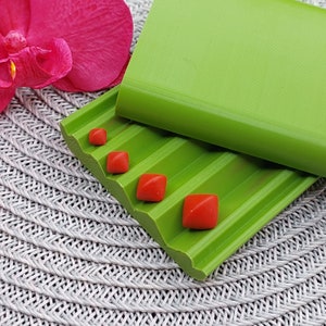 May include: A close-up of a green, ridged soap dish with four small, square, red objects. A solid green rectangular object sits on top of the dish. A pink flower is in the background.