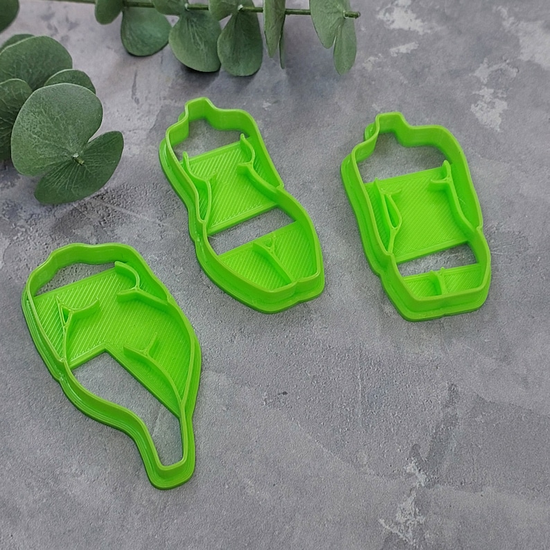 May include: Three bright green plastic cookie cutters in the shape of a bottle and a leaf. The cutters are arranged on a gray surface, with a few green leaves in the background.