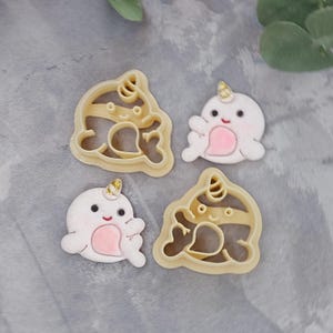 May include: Four cookie cutters shaped like narwhals. Two are solid white with pink accents and two are outlined with a brown stripe. All have gold glitter accents.