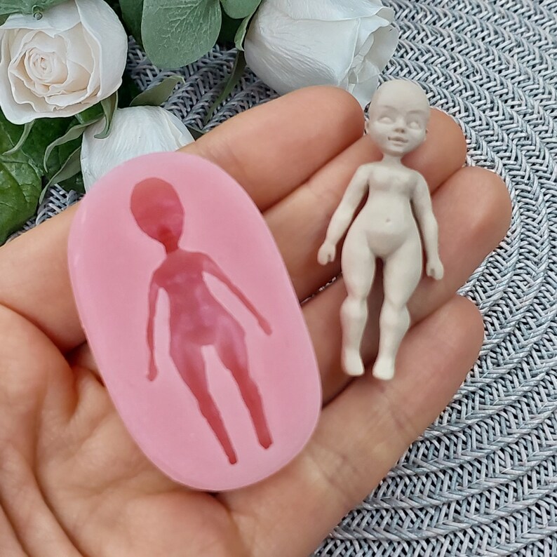 Op de afbeelding: Een roze siliconen mal voor het maken van een klein poppenfiguur. De mal is gevormd als een persoon met een hoofd, armen, benen en een torso. De mal wordt getoond naast een klein, ongeverfd poppenfiguur.