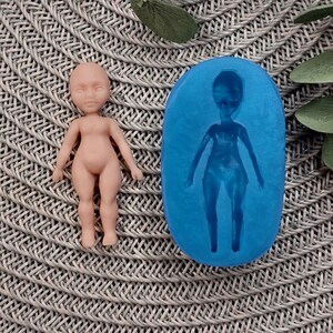 May include: A silicone mould for making a small doll figure. The mould is blue and has a detailed outline of a doll with arms, legs, and a head. A finished doll made from the mould is shown next to it.