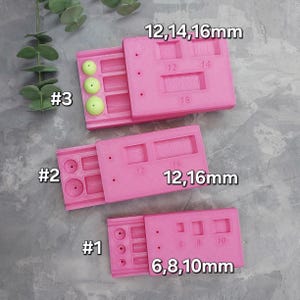 Peut inclure: Trois outils roses en plastique pour la fabrication de bijoux avec des tiroirs coulissants, étiquetés #1, #2 et #3. Chaque outil comporte des trous et des fentes de différentes tailles, avec des mesures en millimètres. L'outil supérieur est étiqueté "12,14,16mm".