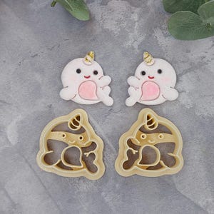 May include: Two white narwhal cookie cutters with gold accents and pink details. The cutters are on a gray background.