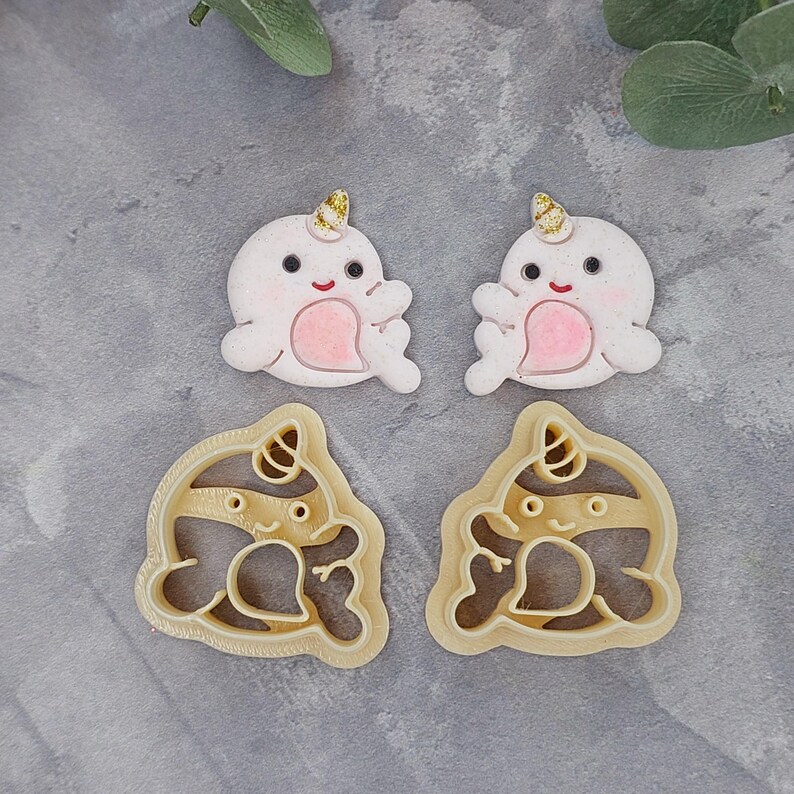 May include: Two white narwhal cookie cutters with gold accents and pink details. The cutters are on a gray background.